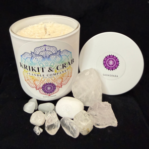 Krikit & Crab Candle Company Sahasrara (Crown Chakra) candle in a white vessel with a purple chakra symbol on the lid. The candle is surrounded by white quartz and clear crystal stones against a black background.