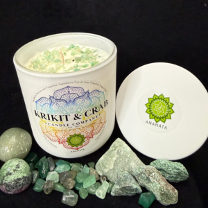 Krikit & Crab Candle Company Anahata (Heart Chakra) candle topped with green crystal chips. The candle is displayed with green aventurine stones and raw green crystals, with the green heart chakra symbol on the lid.