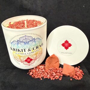 “Krikit & Crab Candle Company Muladhara (Root Chakra) candle featuring red crushed jasper topping. Red jasper stones and raw pieces are displayed beside the candle, with the red root chakra symbol on the lid.”