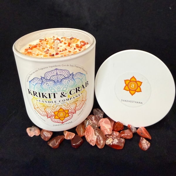 Krikit & Crab Candle Company Svadhisthana (Sacral Chakra) candle topped with orange crystal chips. The candle sits beside polished carnelian stones, with the orange sacral chakra symbol on the lid.