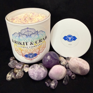 Krikit & Crab Candle Company Ajna (Third Eye Chakra) candle topped with purple amethyst chips. Amethyst tumbled stones and raw pieces are placed in front, with the indigo third eye chakra symbol on the lid.